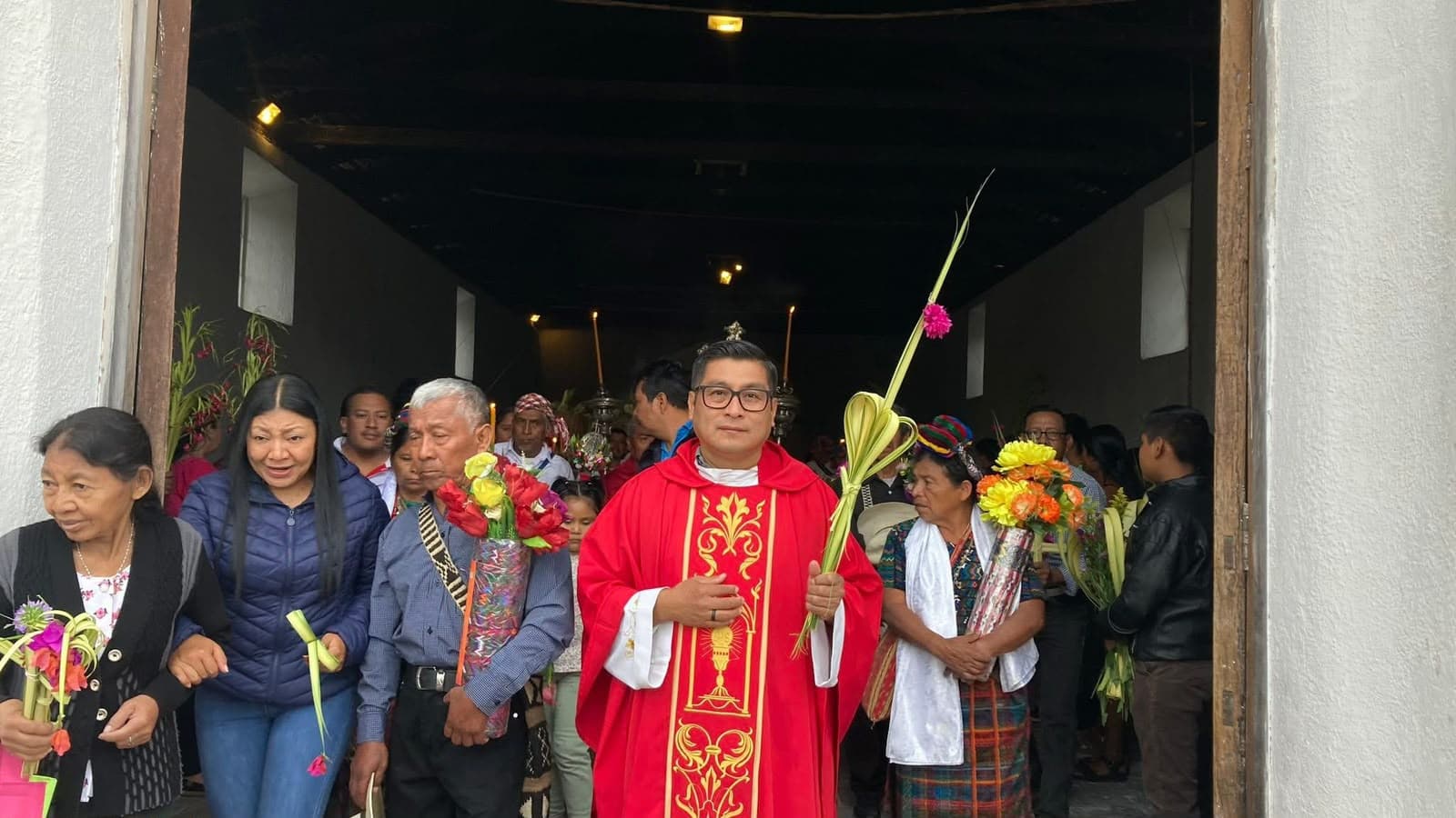 Miembros de la comunidad parroquial junto a un sacerdote, con palmas y flores, a la entrada del templo